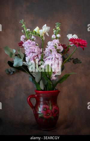 Nature morte verticale d'un bouquet floral avec bouquet rose, freesia blanc et gerbera rouge dans une cruche en céramique rouge peinte à la main sur un fond sombre. Banque D'Images