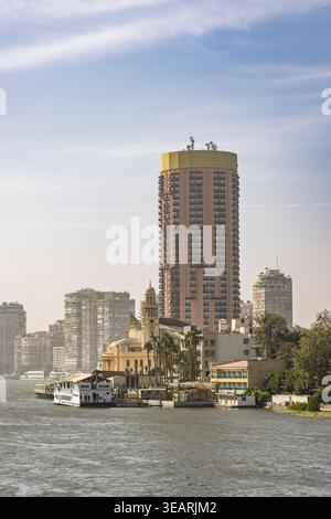Une vue sur la rivière du Caire, en Égypte, avec une grande tour résidentielle, un bateau au bord de la rivière et des bâtiments historiques en bord de mer le long du Nil sous un bleu brumeux Banque D'Images