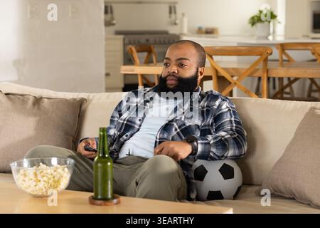 Homme africain assis sur le canapé dans le salon tenant la télécommande TV et ballon de football à côté. Maison, intérieur, open-plan, cuisine, salle à manger, plante, montre-bracelet Banque D'Images