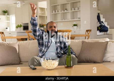 Homme afro-américain d'âge moyen criant et faisant des gestes tout en étant assis sur le canapé avec pop-corn, télécommande. Gentleman, outburst, geste, canapé, open plan, kit Banque D'Images