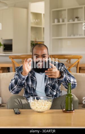 Homme afro-américain en chemise à carreaux assis sur le canapé à la maison, geste, bol de pop-corn, bouteille de bière. Lounge, personne, sourire, geste, open plan, cuisine, Banque D'Images