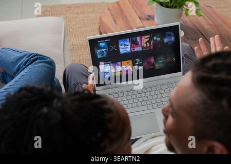 Couple afro-américain assis sur un canapé, parcourant les tuiles sur ordinateur portable, table basse avec plante derrière. Streaming, duo, maison, salon, salon, moderne, minim Banque D'Images