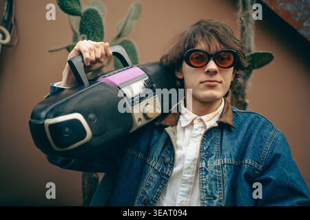Portrait de mode rétro d'un jeune homme élégant avec stéréo portable, lecteur de cassette et CD, lunettes de soleil teintées et veste en Jean, exprimant la culture de la jeunesse, la nostalgie de la musique et le style de vie vintage. Banque D'Images
