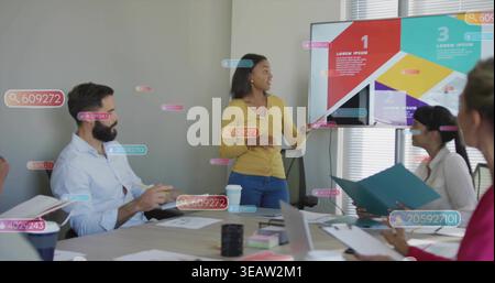 Présentateur gestuel dans un cardigan jaune et un pantalon bleu dirigeant la réunion à table, montrant l'affichage Banque D'Images