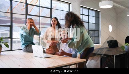 Rires collègues réagissant autour de la table en bois au bureau dans des vêtements de bureau, avec ordinateur portable ouvert Banque D'Images