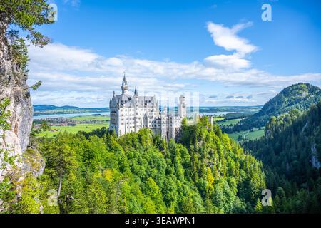 Le château de Neuschwanstein se trouve au sommet d'une colline entourée d'arbres verdoyants. Le ciel est dégagé avec des nuages moelleux. Cet emplacement en Bavière attire de nombreux visiteurs qui admirent la vue. Banque D'Images