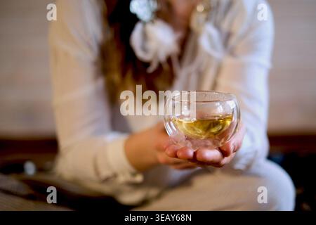 Selective Focus montre une tasse de verre dans la paume ouverte d'une femme, avec sa silhouette assise légèrement floue derrière. Pauses thé et routines de soins personnels Banque D'Images