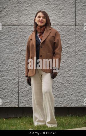 Une femme élégante en vêtements d'extérieur classiques pose près d'un mur texturé gris. La composition reflète le minimalisme, la beauté naturelle et un sens de l'urbain Banque D'Images