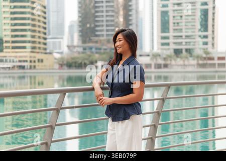 Femme d'affaires asiatique pose près d'un front de mer dans la ville tout en souriant et en profitant de la vue sur les grands bâtiments et l'eau. Banque D'Images
