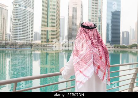 Homme d'affaires arabe se tient sur une rampe au bord de l'eau, regardant la ligne d'horizon des grands bâtiments dans une ville animée. Banque D'Images