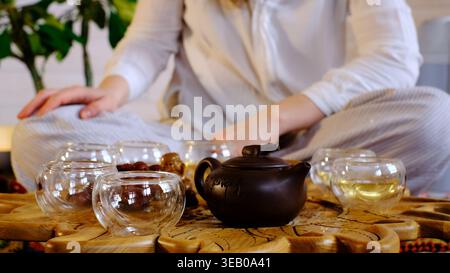 Vue rapprochée de tasses à thé transparentes et d'une théière sombre sur un plateau en bois sculpté tandis qu'une femme est assise floue dans le fond. Les moments de thé attentifs apportent Banque D'Images