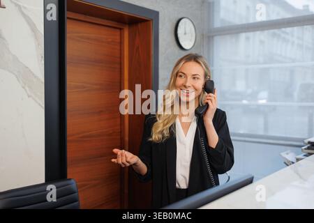 Réceptionniste souriante parlant sur le téléphone de bureau à la réception moderne du bureau Banque D'Images