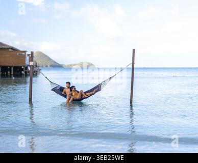 Un couple profite d'un après-midi serein se prélasser ensemble dans un hamac au-dessus des eaux calmes. Un couple multiethnique diversifié d'une femme asiatique et d'un homme caucasien Banque D'Images