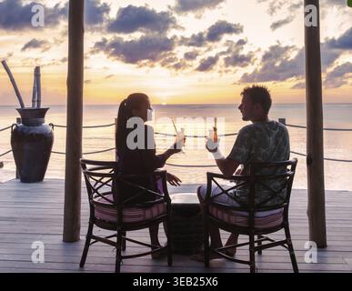 Deux personnes sont assises sur une terrasse en bois, sirotant des cocktails rafraîchissants en regardant le soleil se coucher sur la mer des Caraïbes à Sainte-Lucie. Le ciel est peint avec Banque D'Images