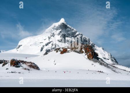 Vue sur un majestueux sommet de montagne enneigé avec une colonie de pingouins et des gens près de drapeaux rouges sur les plaines enneigées par des affleurements rocheux avec lichen orange Banque D'Images