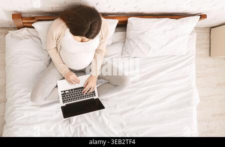 Femme enceinte travaillant sur ordinateur portable tout en étant assise sur le lit dans un cadre de chambre confortable Banque D'Images