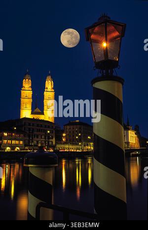 Zurich, Suisse – vue nocturne de la Grossmünster illuminée avec la pleine lune au-dessus et des reflets sur la rivière Limmat. Une scène urbaine sereine avec une architecture historique, des lumières chaudes et une atmosphère calme au bord de la rivière. Banque D'Images