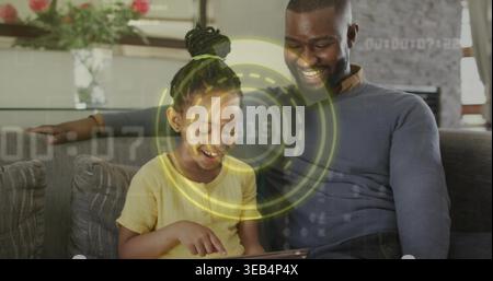 Père souriant penché vers la fille jaune avec bandeau vert à la maison, tapant la tablette, HUD Banque D'Images