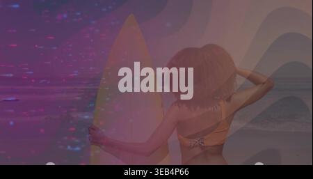 Femme debout en maillot de bain orange tenant une grande planche de surf sur la plage par l'océan, avec recouvrement magenta Banque D'Images