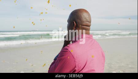 Homme senior debout portant une chemise rose face à l'océan sur des superpositions d'étoiles dorées de sable du rivage, espace de copie Banque D'Images