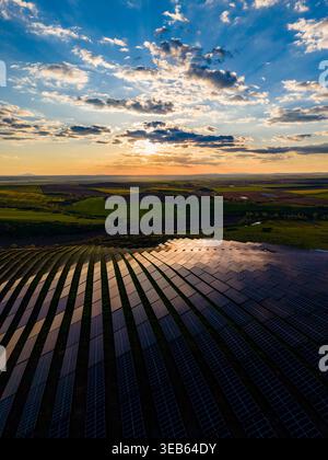 Les panneaux solaires couvrent le sol dans un grand champ alors que le soleil se couche sur l'horizon. Les nuages remplissent le ciel, et le paysage montre des collines ondulantes et des fie ouvertes Banque D'Images