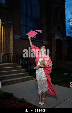 Une personne célèbre la remise des diplômes à l'extérieur d'un bâtiment pendant la nuit. L'individu porte une robe et tient un bonnet tout en se tenant debout sur les marches. Fleurs a Banque D'Images