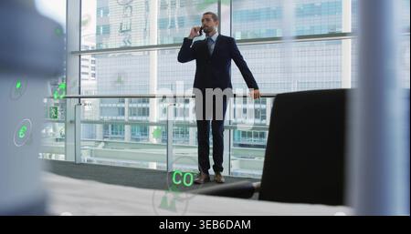 Homme d'affaires parlant portant un costume sombre tenant le téléphone près de la balustrade en verre dans l'atrium, des superpositions AR vertes Banque D'Images