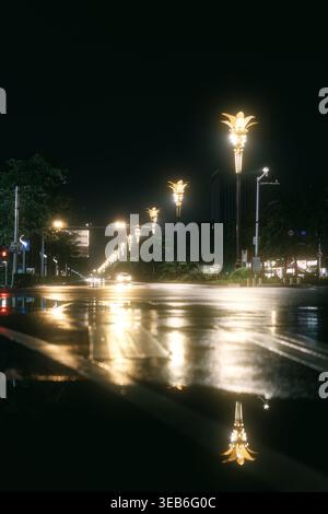 Rue nocturne de la ville avec trottoir humide reflétant des lampadaires ornés et des feux de circulation éloignés Banque D'Images
