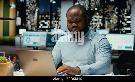 Homme afro-américain fait la pensée critique et la relecture avec des papiers avant de transcrire des données sur ordinateur portable, assis dans le bureau la nuit. Travailleur documentant les idées importantes. Banque D'Images