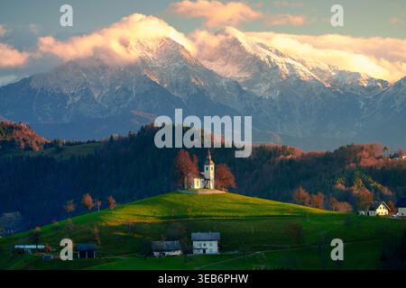 Vue sur l'historique Saint Primus et l'église Felician à Jamnik sur une colline verdoyante avec les Alpes Kamnik-Savinja enneigées sous un ciel doux de coucher de soleil dedans Banque D'Images