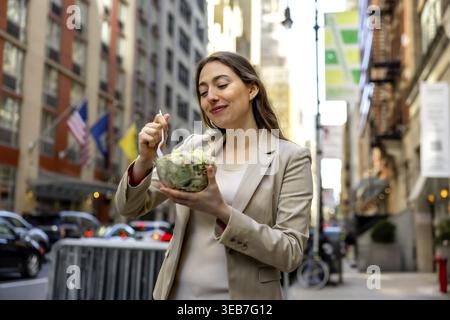 Jeune femme d'affaires en costume dégustant une salade à emporter dans une rue animée de manhattan pendant le déjeuner, montrant un style de vie urbain sain, un équilibre entre vie professionnelle et vie privée Banque D'Images