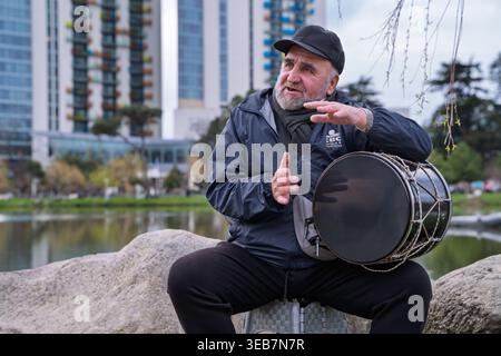 Batoumi, géorgie 21 avril 2026 : homme géorgien âgé jouant un tambour doli et parlant, vêtu de vêtements décontractés, contre le parc urbain de lac backgroun Banque D'Images