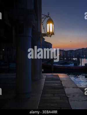 Vue d'une lanterne traditionnelle lumineuse suspendue à une colonne de pierre au crépuscule surplombant le Grand canal avec des bateaux amarrés Venise, Vénétie, Italie. Banque D'Images