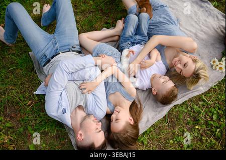 Vue de dessus de la famille heureuse couchée, câlin sur couverture dans le parc d'été. Concept d'environnement sûr, inclusion pour l'enfant avec moulage de bras et lien familial. Parents et enfants se relaxant sur l'herbe pendant le pique-nique Banque D'Images
