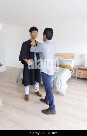 Hommes asiatiques debout dans la chambre, un ajustant le col de robe de graduation noir près du lit Banque D'Images