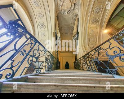 Une vue en contre-plongé montant une grande escalier en marbre avec des rampes en fer forgé noir ornées de motifs en spirale complexes. Le plafond Banque D'Images