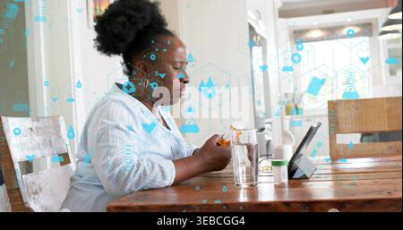 Tenant la bouteille de pilule orange, femme noire portant chemisier vérifiant la tablette à table avec des icônes de santé Banque D'Images