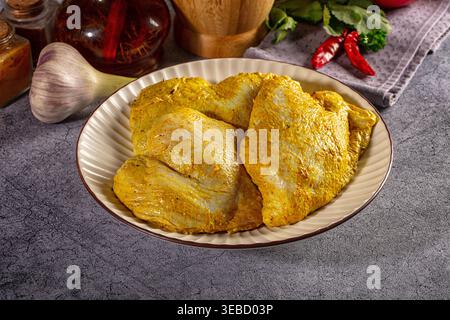 Cuisses de poulet crues marinées dans un mélange de curry jaune et d'épices de curcuma sur une assiette en céramique. Une scène culinaire avec de l'ail frais, des piments et des herbes o Banque D'Images
