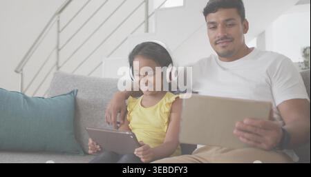 Père assis dans le tee-shirt blanc et fille dans le dessus jaune tenant des tablettes sur le canapé de la maison, écouteurs Banque D'Images