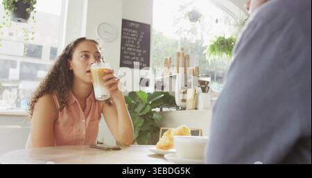 Femme buvant dans le haut de saumon sans manches sirotant un latte glacé à la table de la fenêtre du café, avec croissant Banque D'Images