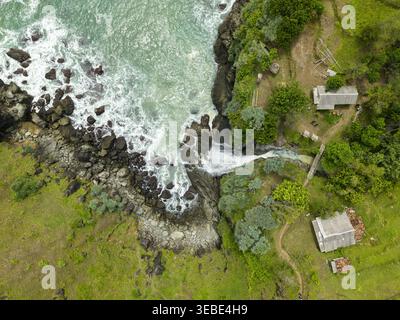 Vue aérienne de la chute d'eau de Banyutibo cascadant au-dessus des rochers côtiers sombres dans les vagues turquoise de l'océan entouré de falaises verdoyantes et de petits b ruraux Banque D'Images