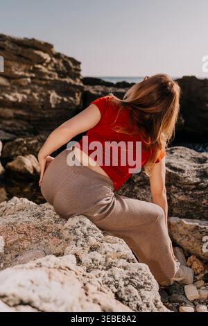 Femme rochers de mer assis sur des pierres côtières portant une chemise rouge et un pantalon brun pendant le coucher du soleil avec la lumière de l'heure dorée Banque D'Images