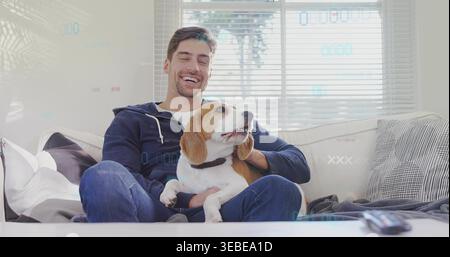Homme souriant tenant beagle avec col sur canapé dans le salon, portant un sweat à capuche bleu marine et un Jean Banque D'Images