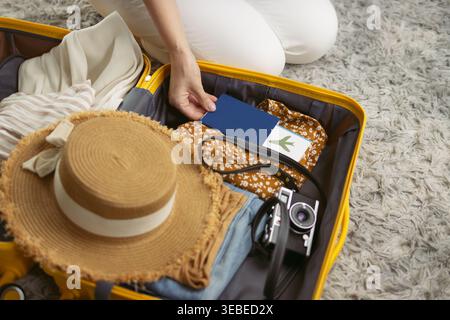 Femme emballant une valise jaune pour les vacances d'été, plaçant passeport et carte d'embarquement à côté des vêtements, chapeau de paille et appareil photo dans une vue de dessus qui Banque D'Images
