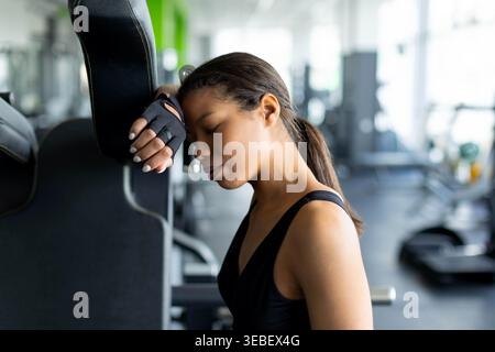 Jeune femme noire épuisée penchant la tête contre la machine de gymnastique avec les yeux fermés Banque D'Images