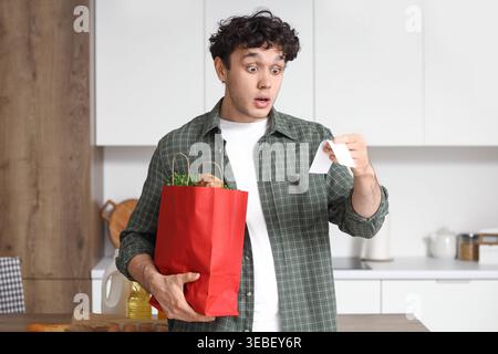 Jeune homme choqué avec sac de nourriture et réception dans la cuisine. Concept d'augmentation des prix Banque D'Images