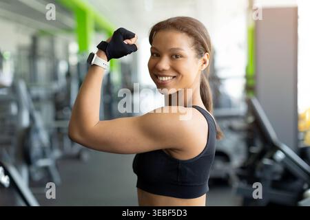 Femme noire heureuse fléchissant le muscle biceps et souriant à la caméra dans la salle de gym Banque D'Images