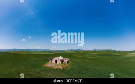Vue aérienne d'une ferme en pierre abandonnée entourée de vastes champs de blé vert et de collines ondulantes sous un ciel bleu clair Pouilles, Italie. Banque D'Images