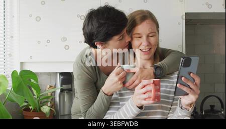 Femme penchée dans le haut d'olive embrassant partenaire dans le haut rayé, tenant des tasses, téléphone dans la cuisine à la maison Banque D'Images