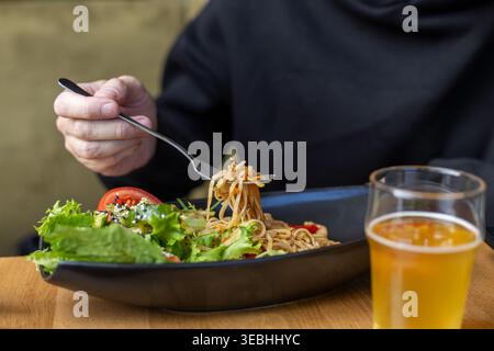 Homme mangeant des nouilles asiatiques avec de la bière. Gros plan d'un homme mangeant des nouilles asiatiques accompagnées de salade fraîche et de bière, montrant un dîner décontracté dans un restaurant. Banque D'Images
